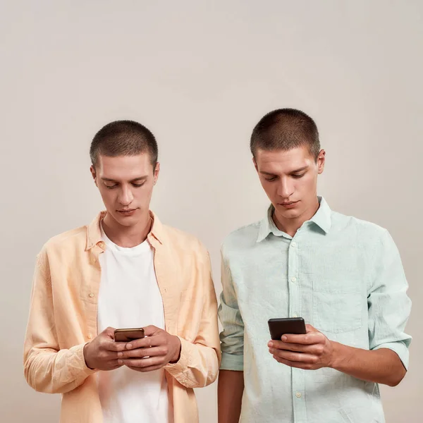 Due giovani fratelli gemelli caucasici in abbigliamento casual utilizzando smartphone mentre in piedi isolati su sfondo beige — Foto Stock