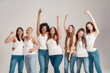 Beyaz gömlek ve kot pantolon giyen, neşeli görünen, birlikte poz veren bir grup güzel genç kadın gri arka planda izole edilmiş.