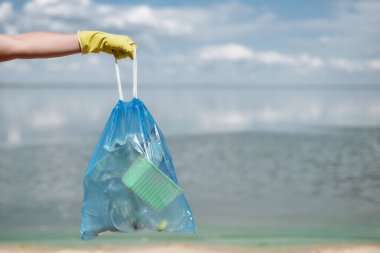 Gönüllüler, plaj plastik atıkları ve diğer çöpleri temizlerken çöp torbalarını denize ve gökyüzüne karşı tutan sarı lastik eldiven veriyorlar.