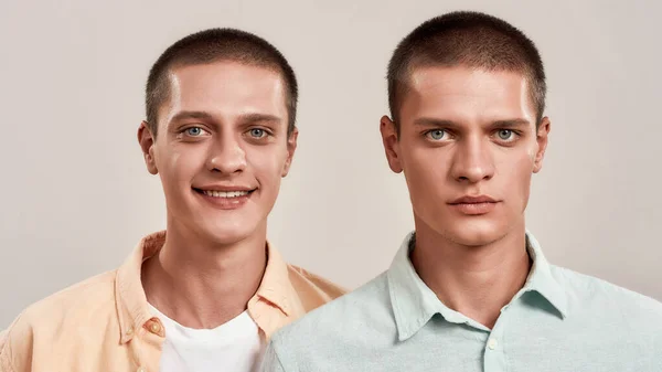 Close up retrato de dois jovens irmãos gêmeos caucasianos posando juntos em estúdio, olhando para a câmera com diferentes expressões faciais enquanto em pé isolado sobre fundo bege — Fotografia de Stock