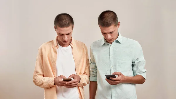 Due giovani fratelli gemelli caucasici in abbigliamento casual utilizzando smartphone mentre in piedi isolati su sfondo beige — Foto Stock