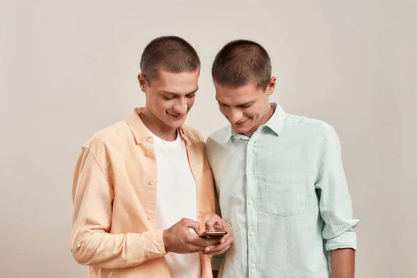 Dois jovens irmãos gêmeos alegres em uso casual usando o aplicativo do smartphone enquanto estão juntos isolados sobre o fundo bege — Fotografia de Stock