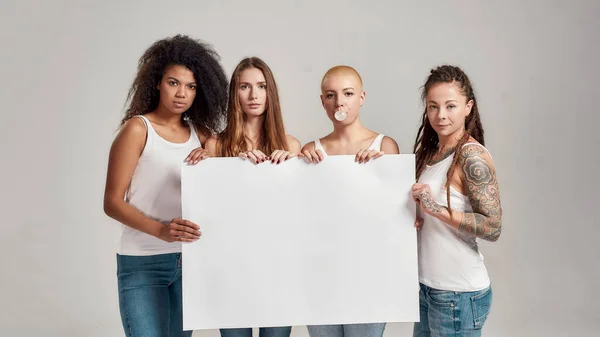 Cuatro mujeres diversas mirando a la cámara mientras sostienen, de pie con pancarta en blanco en sus manos aisladas sobre un fondo gris. Concepto de feminismo e igualdad —  Fotos de Stock