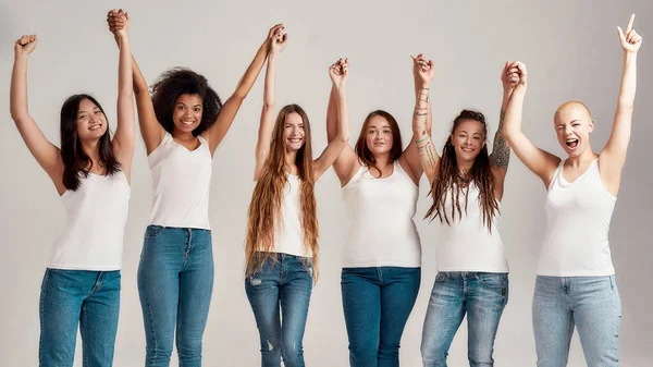 Grupa pięciu pięknych, różnorodnych młodych kobiet ubranych w białą koszulę i dżinsy jeansowe podnoszące ramiona, patrząc wesoło przed kamerą, pozując razem na szarym tle — Zdjęcie stockowe