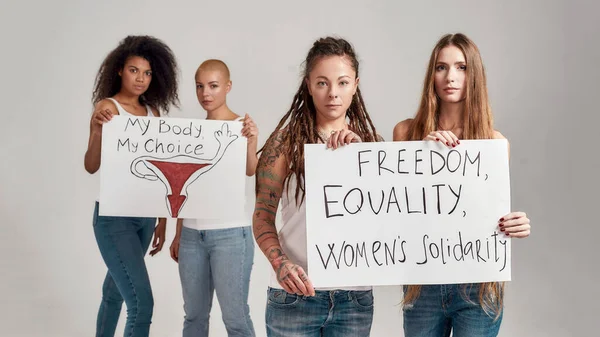 Quattro donne diverse che guardano la macchina fotografica mentre tengono, in piedi con striscioni tra le mani isolati su sfondo grigio. Concetto di femminismo e uguaglianza — Foto Stock