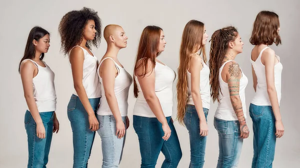 Sekelompok wanita muda cantik yang beragam mengenakan kemeja putih dan celana jeans denim melihat ke samping sambil berpose, berdiri terisolasi di atas latar belakang abu-abu — Stok Foto