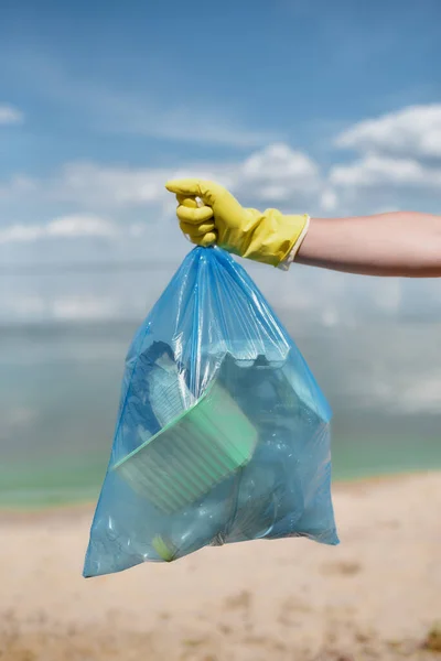 Voluntario o eco activista con guantes de goma amarillos sosteniendo la bolsa de basura con residuos de plástico y otra basura contra el agua y el cielo mientras limpia la playa — Foto de Stock