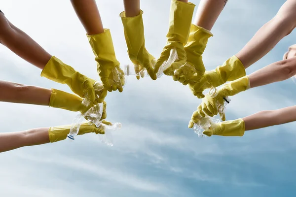 Städa upp dagen. Grupp frivilliga eller miljöaktivister bär gula skyddshandskar gummi som håller plastflaskor mot blå himmel. — Stockfoto