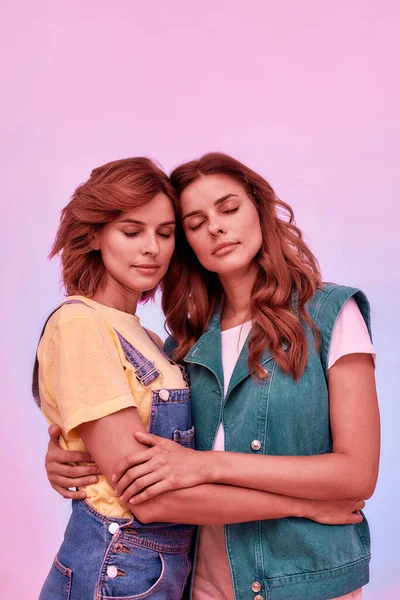 Portrait of two attractive young girls, twin sisters holding each other, posing together with eyes closed isolated over pink background
