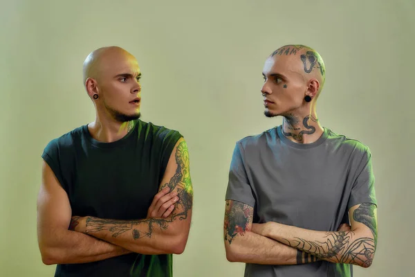 Horizontale Aufnahme von zwei jungen kaukasischen Zwillingsbrüdern mit Tattoos und Piercings, die die Arme verschränkt halten und einander beim gemeinsamen Posieren im Studio anschauen — Stockfoto