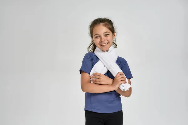 Маленькая спортивная девочка в спортивной одежде улыбается в камеру, держа полотенце вокруг шеи, стоя изолированно на белом фоне — стоковое фото