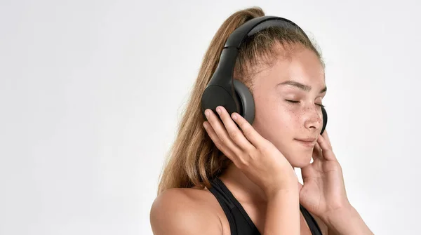 Retrato de una linda niña deportiva con auriculares, escuchando música, de pie con los ojos cerrados mientras posa aislada sobre un fondo blanco — Foto de Stock
