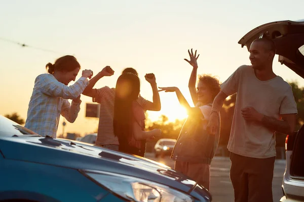 Een groep jonge vrouwen en mannen die dansen op muziek en plezier hebben buiten op een parkeerplaats bij hun auto 's — Stockfoto