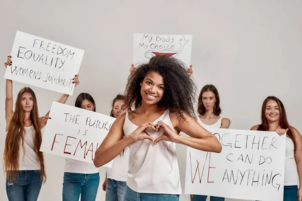 Giovane donna afroamericana in camicia bianca che mostra cuore o segno d'amore, sorridendo alla macchina fotografica. Gruppo di diverse donne con striscioni di protesta per il potere e i diritti delle donne sullo sfondo — Foto Stock