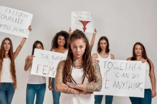 Ung kaukasisk tatuerad kvinna med dreadlocks som ser allvarlig ut. Grupp av olika kvinnor som håller protestbanners för kvinnlig makt och rättigheter i bakgrunden — Stockfoto