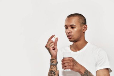 İlaç alıyorum. Koyu tenli, dövmeli bir adam elinde bir bardak suyla beyaz bir arka planda duran bir hapa bakıyor. 