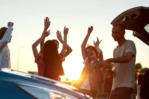 A group of young well-dressed friends dancing to music having a good time together outside on a parking site near their cars