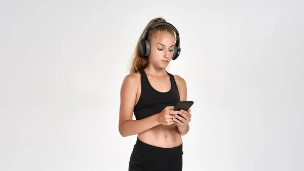 Retrato de una linda niña deportiva que usa auriculares, usando un teléfono inteligente mientras posa aislado sobre un fondo blanco — Foto de Stock