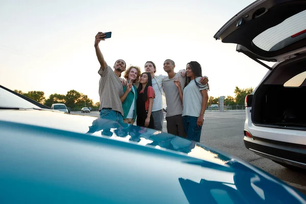 Zes jonge vrijetijds geklede vrienden die een leuke tijd samen hebben, elkaar glimlachend knuffelen en buiten een groepsfoto maken op een parkeerplaats bij hun auto 's. — Stockfoto