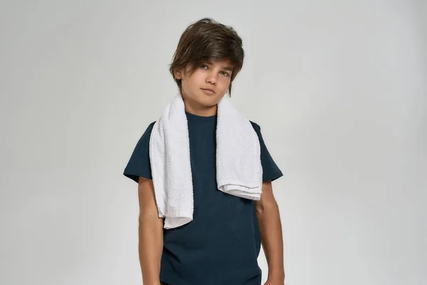 Μικρό σπορ αγόρι με αθλητικά ρούχα κοιτάζοντας την κάμερα, στέκεται με λευκή πετσέτα γύρω από το λαιμό του απομονωμένο σε λευκό φόντο — Φωτογραφία Αρχείου