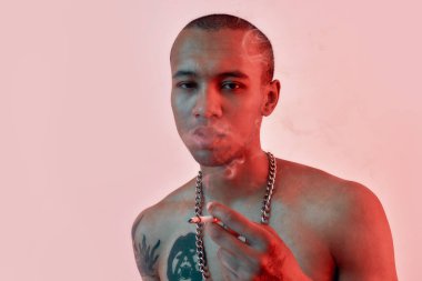 Sigara içiyorum. Siyah tenli, üstsüz, dövmeli sigara içerken duman çıkaran ve kameraya bakan bir erkek.