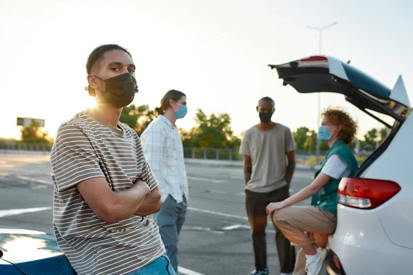 Κοντινό πλάνο ενός σκουρόχρωμου Λατίνου που φοράει μάσκα κοιτάζοντας μια κάμερα με τα χέρια σταυρωμένα δίπλα σε ένα αυτοκίνητο έξω σε ένα χώρο στάθμευσης με τους φίλους του σε φόντο — Φωτογραφία Αρχείου
