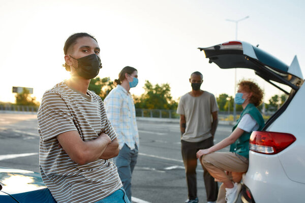 A close up of a dark-skinned latina guy wearing a mask looking into a camera with hands crossed next to a car outside on a parking site with his friends on a background