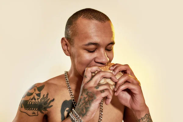 Comer comida. Um homem tatuado de pele escura segurando um hambúrguer com duas mãos mordendo-o com os olhos fechados — Fotografia de Stock