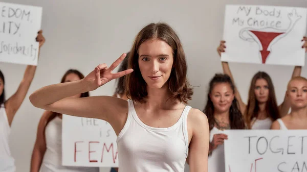 Beautiful young caucasian woman with short hair showing peace sign, smiling at camera. Group of diverse women holding protest banners in the background — Stock Fotó