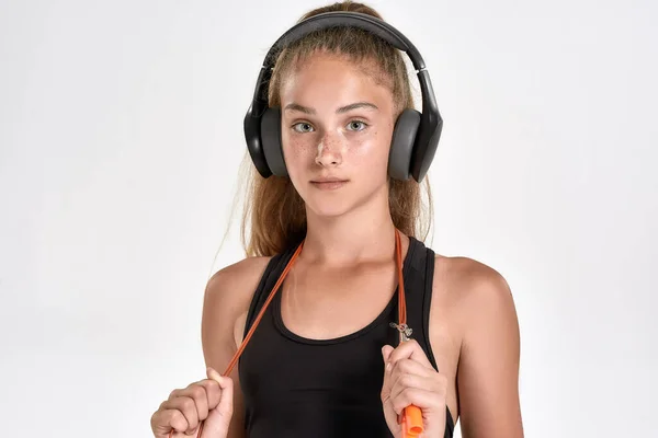 Primer plano retrato de niña linda deportiva en los auriculares mirando a la cámara, de pie con cuerda de salto mientras posa aislado sobre fondo blanco — Foto de Stock