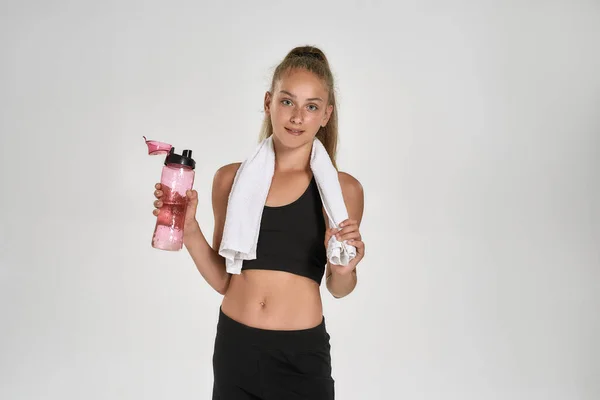 Porträt eines niedlichen sportlichen Mädchens in schwarzer Sportbekleidung, das in die Kamera blickt und eine Wasserflasche hält, während es mit einem Handtuch um den Hals vor weißem Hintergrund isoliert steht — Stockfoto