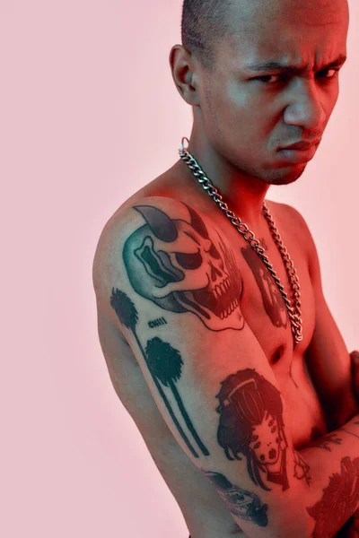 Tatuagens a aparecer. Homem de pele escura com tatuagens em pé lateralmente a uma câmera topless com as mãos cruzadas no peito e expressão facial descontente — Fotografia de Stock