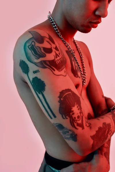 Tattoos showing.A dark-skinned man with tattoos standing sideways to a camera topless, wearing a large link chain slightly embracing himself — Photo
