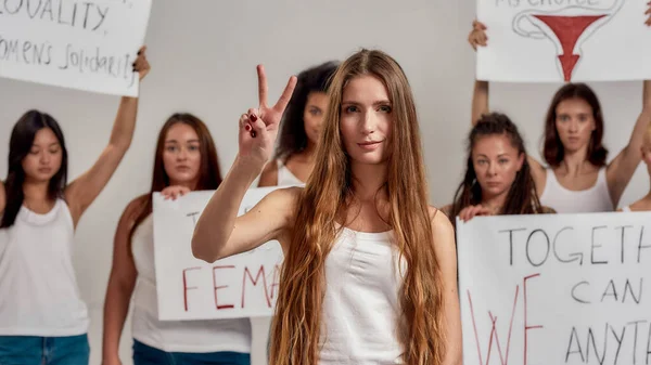 Young caucasian woman with long hair showing peace sign, looking at camera. Group of diverse women holding protest banners in the background — Stock Fotó