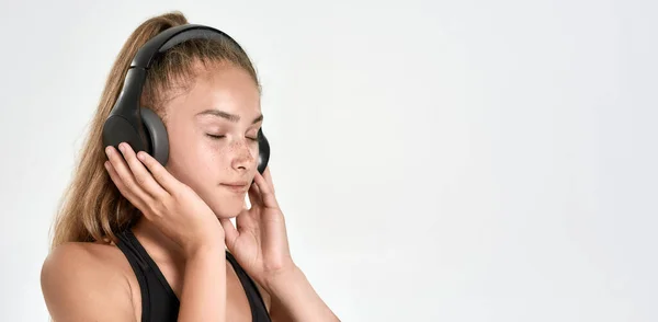 Retrato de una linda niña deportiva con auriculares, escuchando música, de pie con los ojos cerrados mientras posa aislada sobre un fondo blanco — Foto de Stock