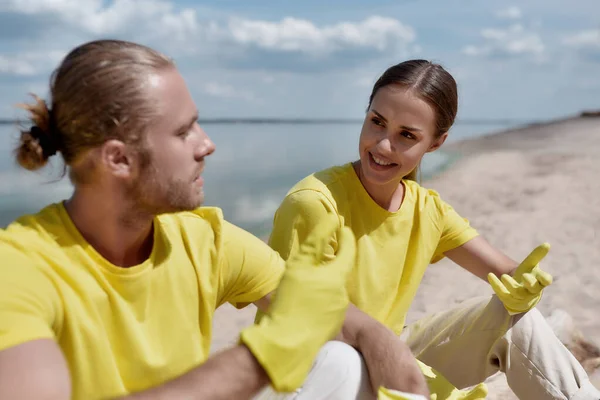Two young happy eco activists wearing uniform and rubber gloves resting after cleaning a beach and discussing something — Photo