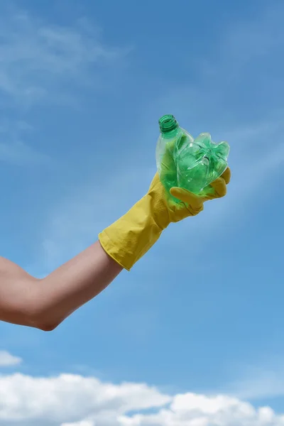 Proteger nuestro medio ambiente. Tiro vertical de un voluntario con guante de basura amarillo sosteniendo botella de plástico usado contra el cielo azul — Foto de Stock