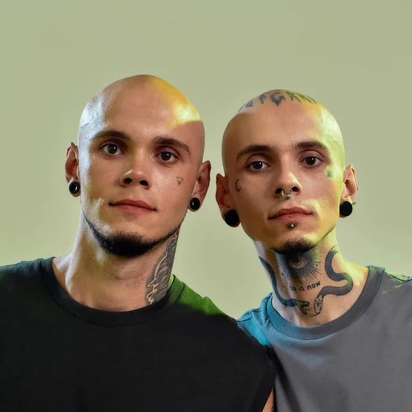 两名年轻男子的近照，他们是双胞胎兄弟，身上有纹身和穿孔，看着相机，他们站在一起，在浅绿色的背景上，彼此隔离 — 图库照片