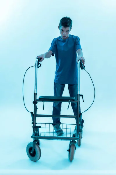 Full length shot of teenaged disabled boy with cerebral palsy looking at camera, taking steps with his walker isolated over blue background with neon light — Stock Photo, Image