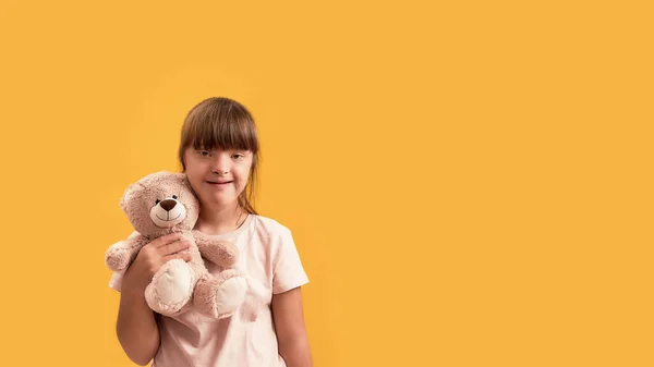 Retrato de una chica discapacitada con síndrome de Down sonriendo a la cámara, sosteniendo su osito de peluche mientras posa aislada sobre un fondo amarillo —  Fotos de Stock