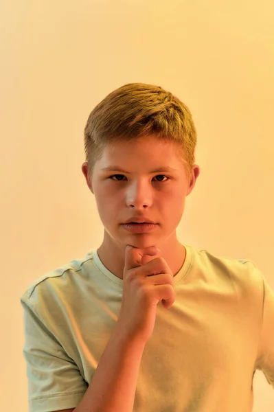 Portrait of thoughtful disabled boy with Down syndrome looking at camera, holding hand on chin while posing isolated over yellow light background — Stock Photo, Image