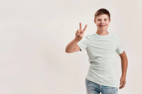 Lycklig funktionshindrade pojke med Down syndrom ler mot kameran, visar fred tecken med ena handen när du står isolerad över vit bakgrund — Stockfoto