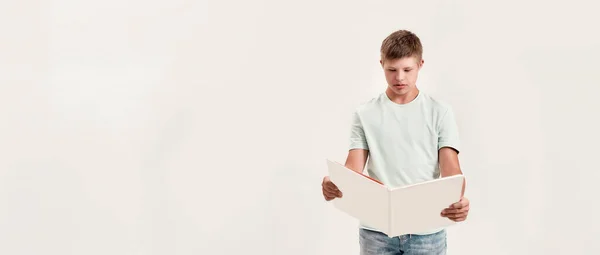 Teenaged disabled boy with Down syndrome looking focused while reading a book, standing isolated over white background — Stock Photo, Image