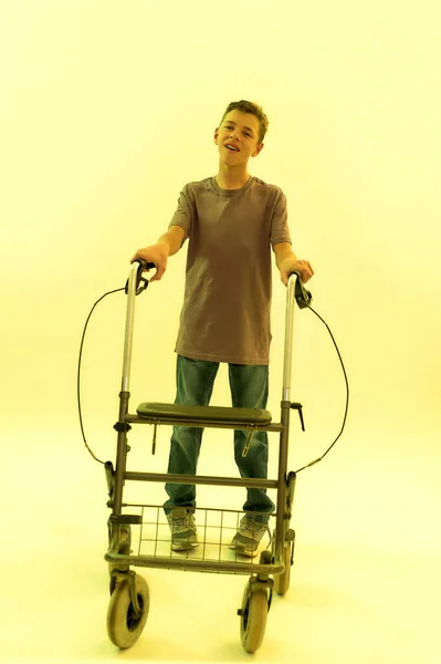 Full length shot of cheerful teenaged disabled boy with cerebral palsy looking at camera, taking steps with his walker isolated over yellow light background — Stock Photo, Image