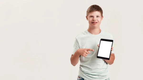 Niño discapacitado feliz con síndrome de Down sonriendo, sosteniendo y apuntando a la tableta PC con pantalla en blanco mientras está de pie aislado sobre fondo blanco —  Fotos de Stock