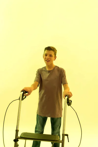 Cheerful teenaged disabled boy with cerebral palsy smiling at camera, taking steps with his walker isolated over yellow light background — Stock Photo, Image