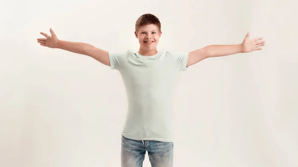 Счастливый мальчик-инвалид с синдромом Дауна улыбается в камеру во время позирования, стоя с широко раскрытыми руками, растянутыми на белом фоне — стоковое фото