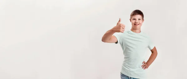 Joyful disabled boy with Down syndrome smiling at camera, showing thumbs up with one hand while standing isolated over white background — Photo