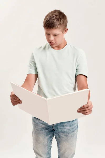 Garçon handicapé adolescent avec trisomie 21 regardant concentré tout en lisant un livre, debout isolé sur fond blanc — Photo