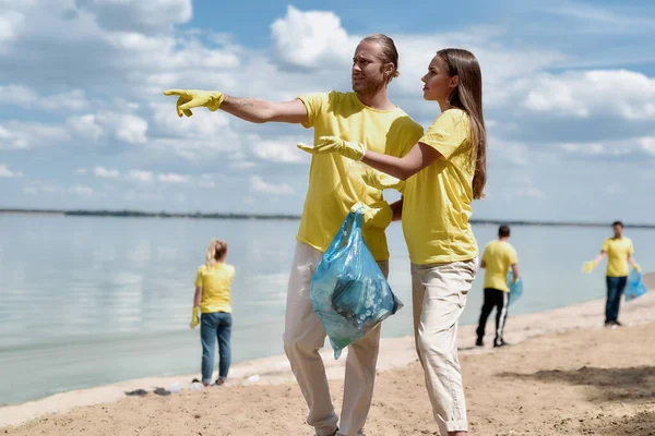 Activistas ambientales. Dos hombres y mujeres jóvenes sosteniendo la bolsa de basura y discutiendo algo mientras limpian el área de la playa de plástico con el equipo de voluntarios — Foto de Stock